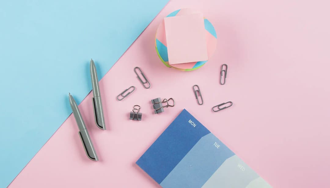 Stifte, Büroklammern und Haftnotizen auf hellblau-rosa Hintergrund