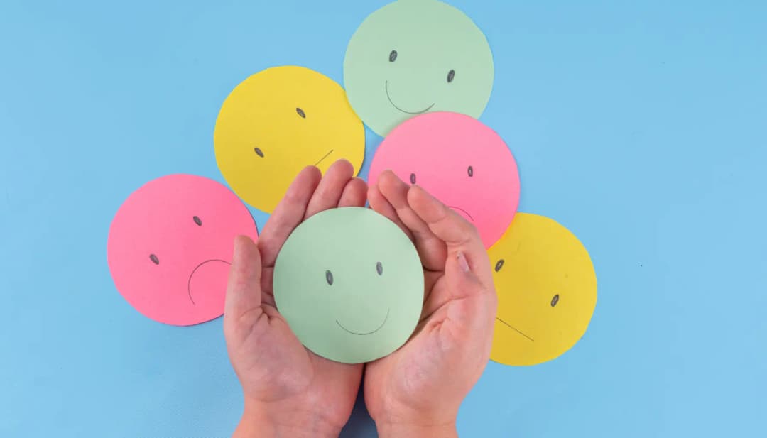 Hände halten einen lächelnden Smiley zwischen bunten Gesichtern aus Papier