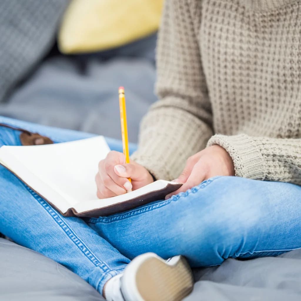 Person im Strickpullover schreibt mit Bleistift in ein Notizbuch auf dem Schoß