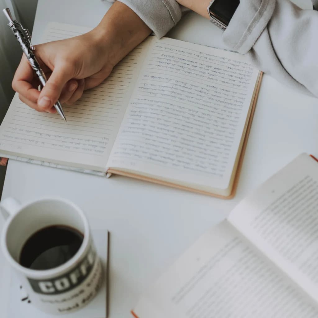 Hand schreibt in ein Notizbuch, daneben eine Tasse Kaffee