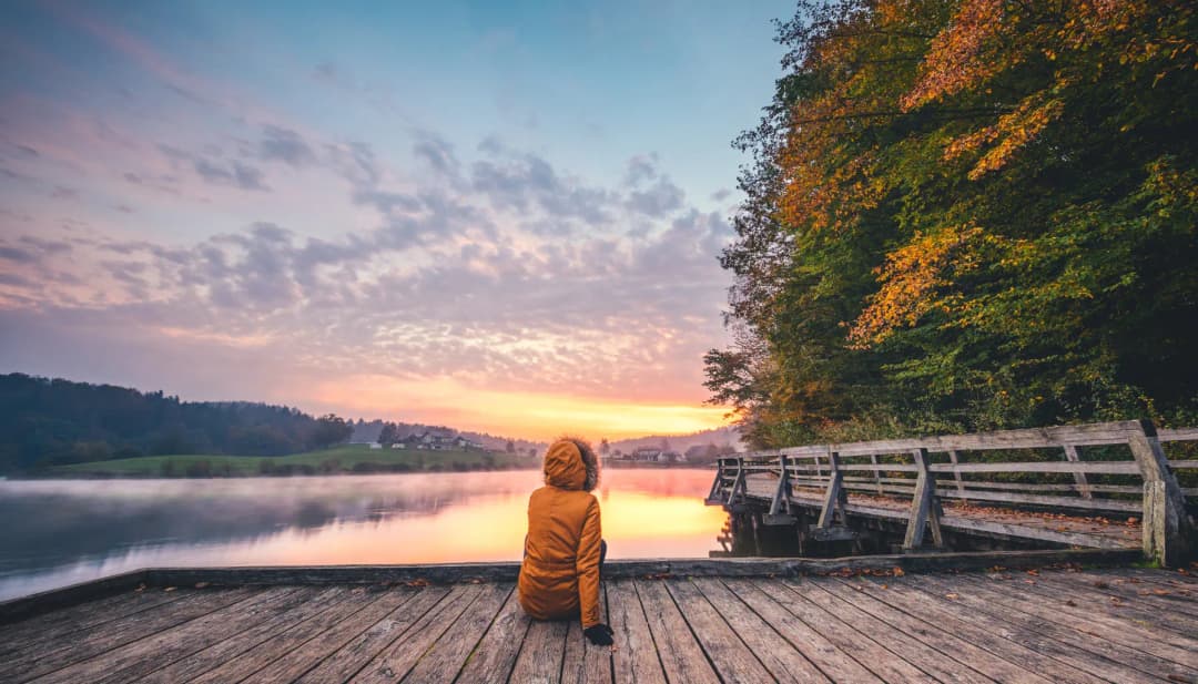 Person sitzt bei Sonnenaufgang auf einem Steg am ruhigen See