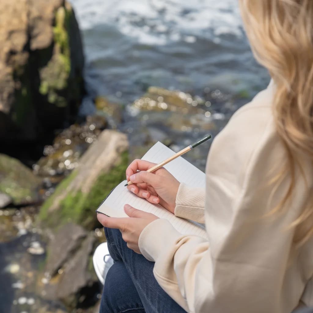 Frau sitzt auf Felsen am Meer und schreibt in ein Notizbuch