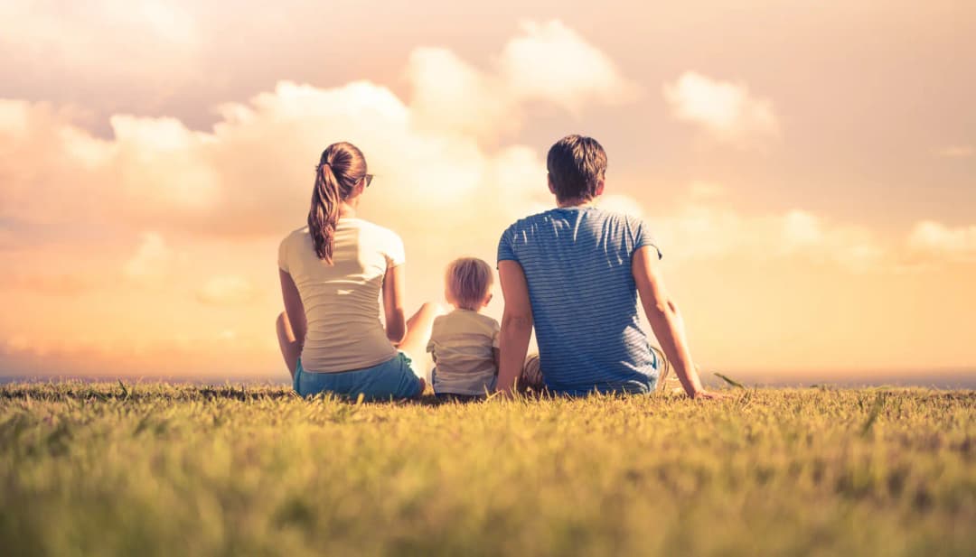 Familie mit Kind sitzt von hinten gesehen auf einer Wiese im Abendlicht