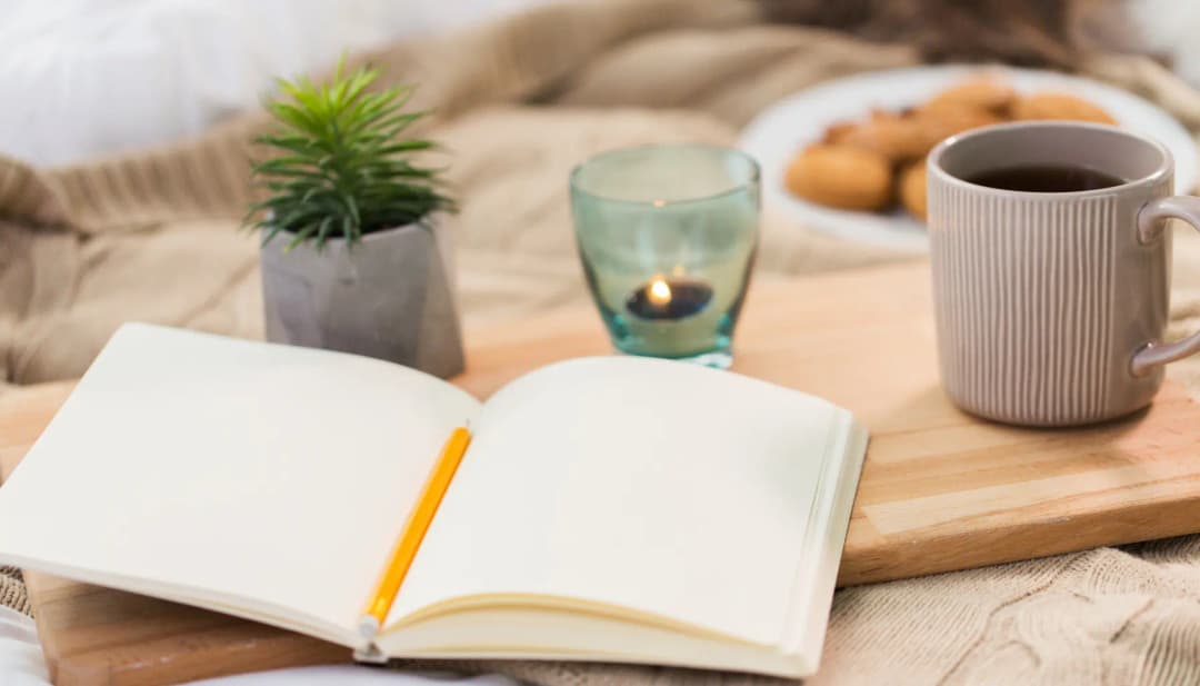 Aufgeschlagenes Notizbuch mit Stift, Tee, Kerze und Keksen auf einem Tablett