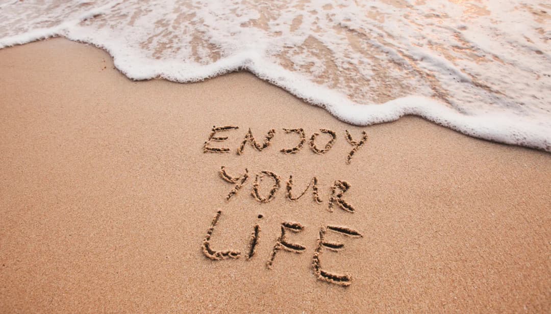 Schriftzug „Enjoy your life“ im Sand, eine Welle läuft heran