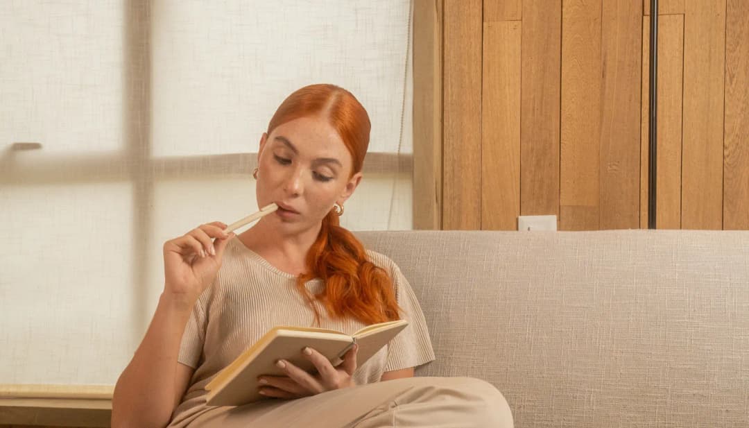 Rothaarige Frau sitzt mit Buch auf dem Sofa und denkt nach
